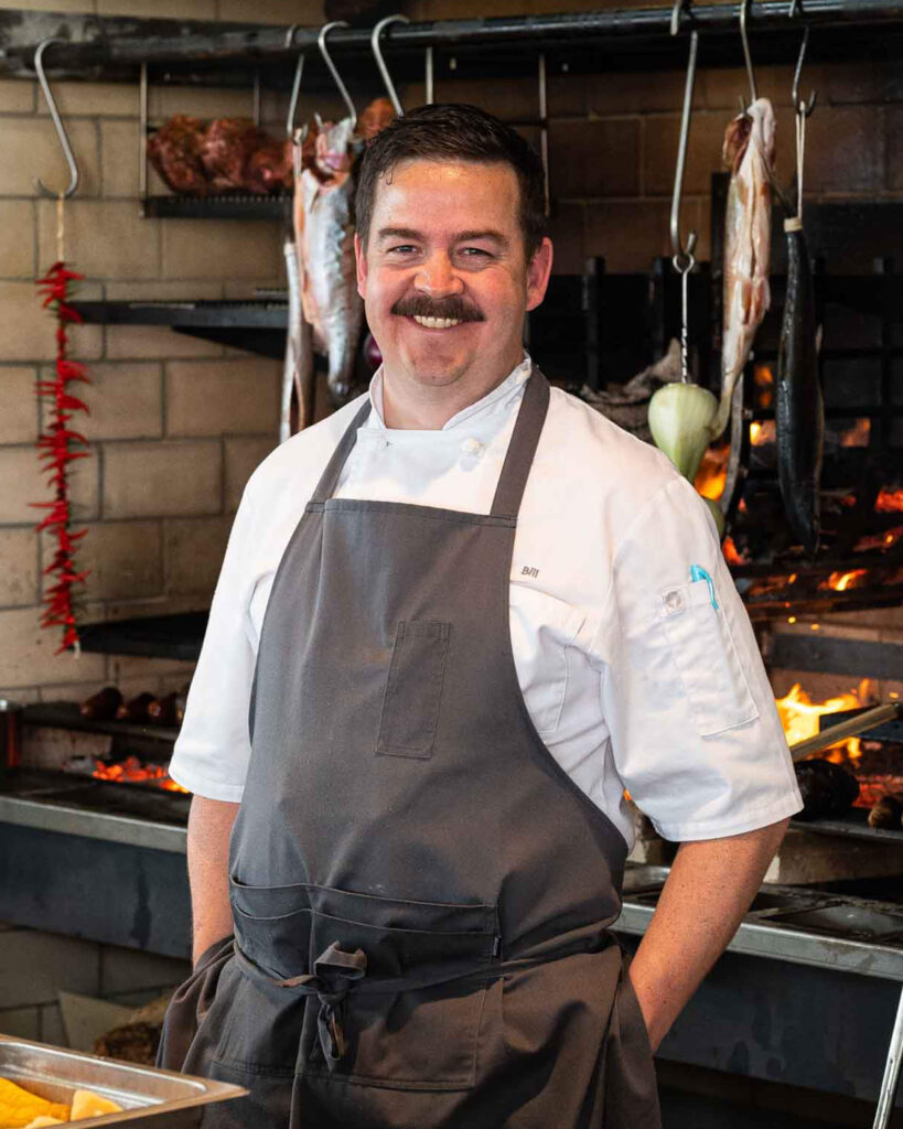 Bill Osborne DISTRICT EXECUTIVE CHEF smiles poses in front of a live fire cooking station