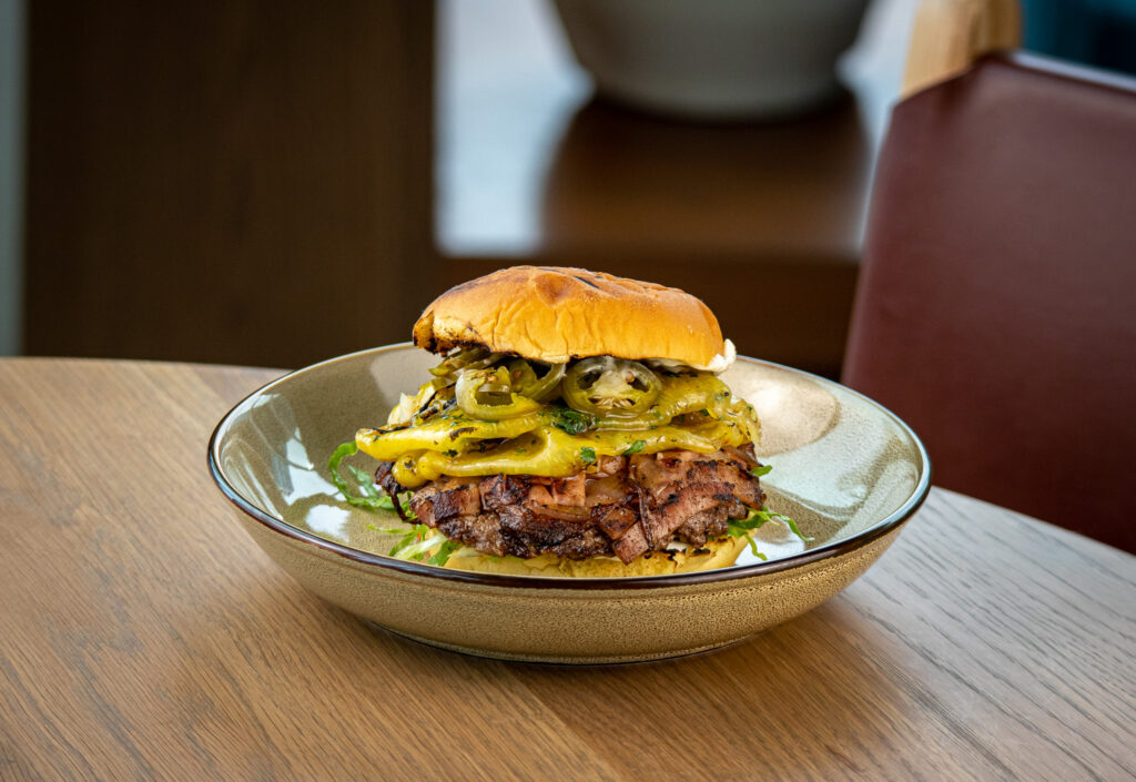 Em-Bologna Burger with juicy beef chuck and bologna smash patty, with toppings of jalapeño aioli, shredded iceberg lettuce, and wood-fired miso pineapple.
